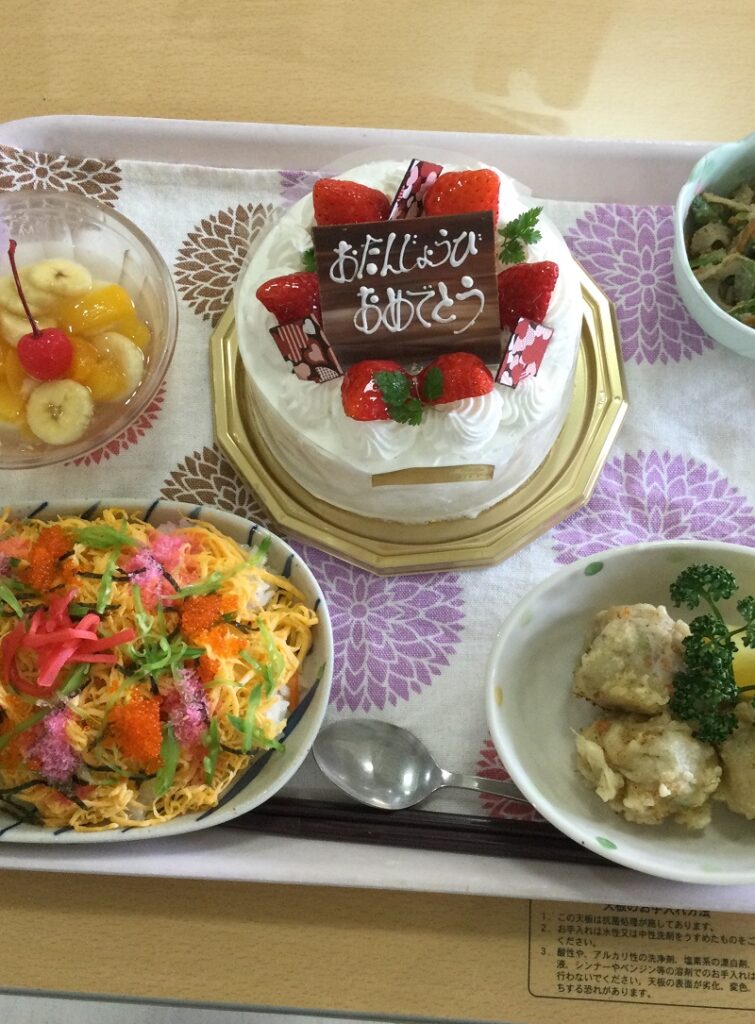 誕生日祝いの食事。ケーキと複数のお祝い料理が並んでいる施設の行事風景。